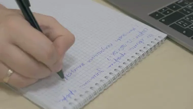 Close-up of a hand writing notes in a notebook beside a laptop.